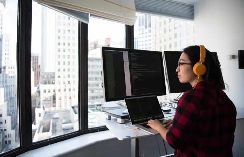 Mujer programando en frente de una ventana, utilizando varias pantallas.