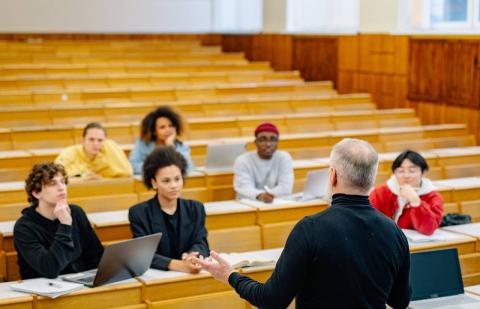 Alumnos sentados y escribiendo en cuadernos en clase mientras el profesor explica