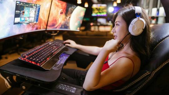 Una gamer, reclinada, juega a videojuegos en su ordenador con tres pantallas.