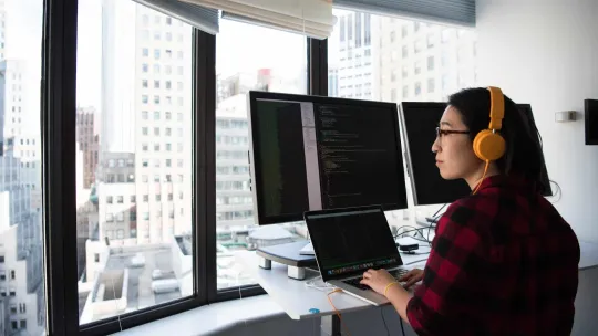 Mujer programando en frente de una ventana, utilizando varias pantallas.