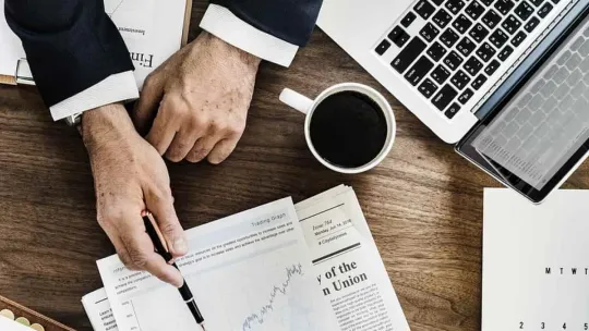 Un técnico en estudios de mercado expone su análisis con documentos y un portátil en una oficina. 