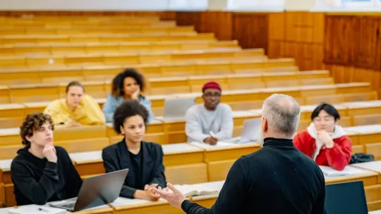 Alumnos sentados y escribiendo en cuadernos en clase mientras el profesor explica